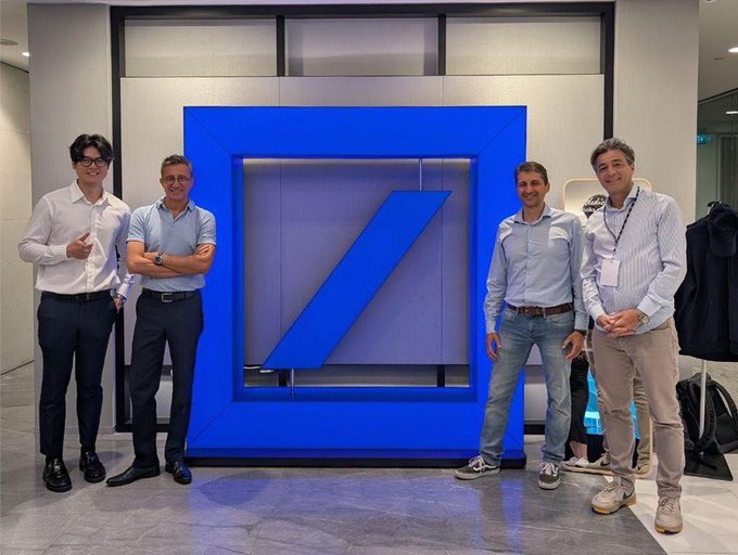 Four men posing for a photo in front of a large Deutsche Bank logo, dressed in business casual attire.