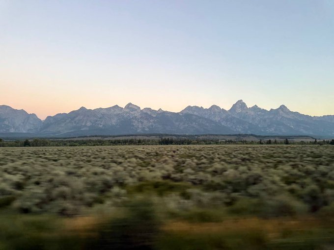 A scenic landscape photograph of a snow-capped mountain range, likely the Grand Tetons, viewed across a sagebrush plain at dawn or dusk.