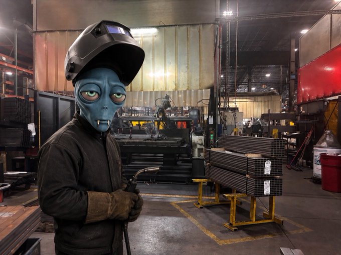 A figure wearing a welding helmet and a large alien mask holds a welding torch in an industrial workshop setting.