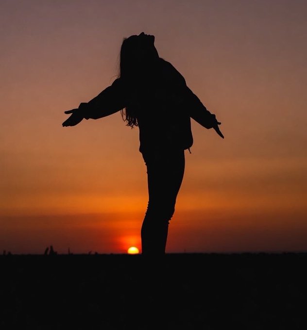 A silhouette of a woman standing with her arms outstretched at sunset, with the sun setting directly behind her on the horizon.