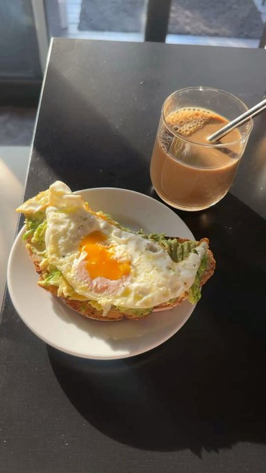 A breakfast scene with a fried egg on avocado toast next to a glass of coffee with milk on a dark table.