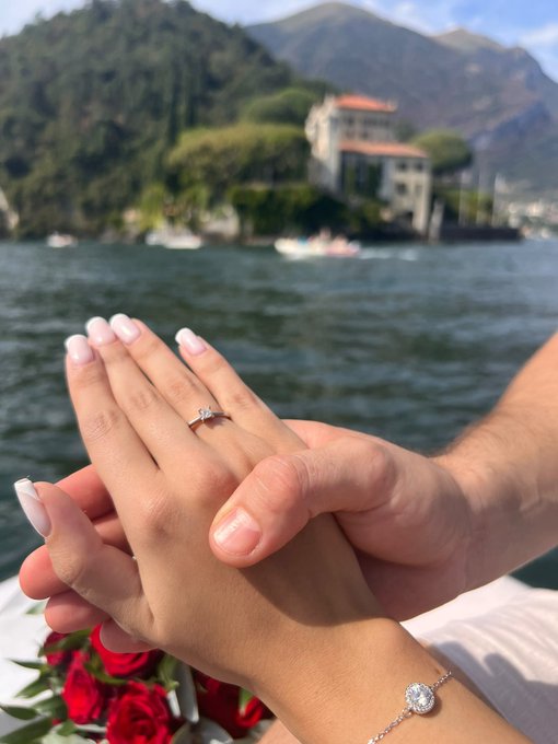 A close-up of a woman's hand with a French manicure and diamond engagement ring resting on a man's hand, with a diamond bracelet visible and red roses in the foreground against a scenic lakeside background.