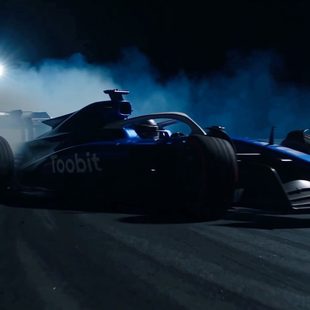 A dark, cinematic image shows a blue Formula 1 race car with 'Toobit' printed on the side, surrounded by smoke or fog under a dramatic blue light.