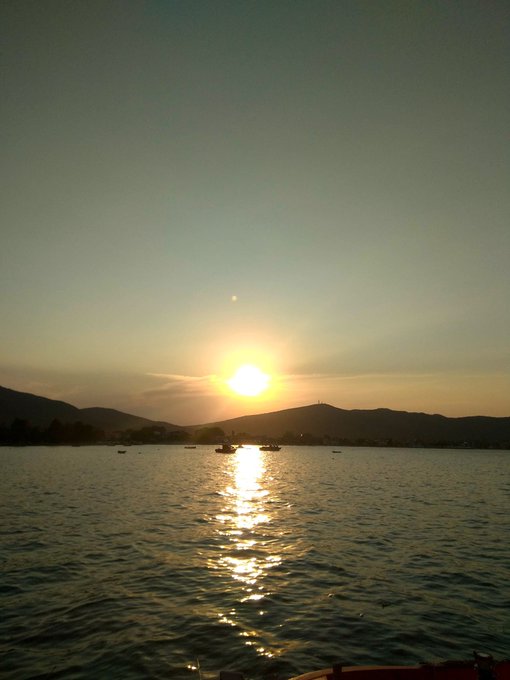 A serene sunset scene over a calm body of water, with the sun's reflection creating a golden path on the surface and silhouetted mountains in the distance.