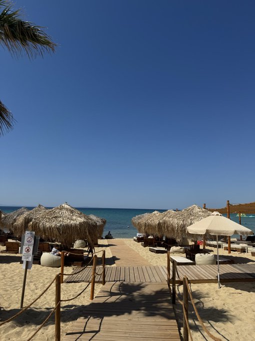 A wooden boardwalk leads to a pristine beach lined with thatched sun umbrellas and a white parasol under a clear blue sky, with a visible sign reading "NO SWIM HERE NO DIVING NO JUMPING" and "EUROMMAXX" posted near the entrance. The serene seaside setting perfectly matches the relaxed vibe of a well-earned 2-week vacation, while the "EUROMMAXX" text directly references the user's "euromaxxing" post in connection with Hyperliquid.