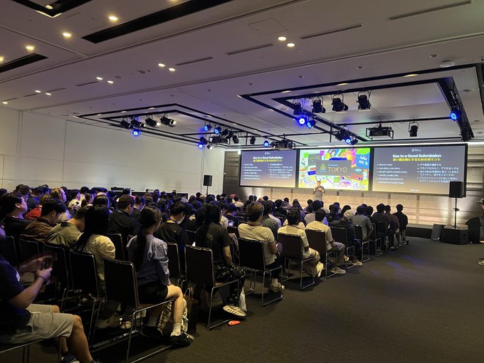 A large audience seated in a conference hall watching a presentation on stage with screens displaying 'How to be a Good Refurbisher'.