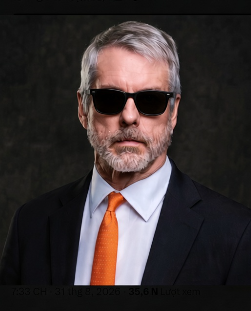 A man with gray hair and beard wearing sunglasses, a black suit, white shirt, and orange tie stands against a dark background.