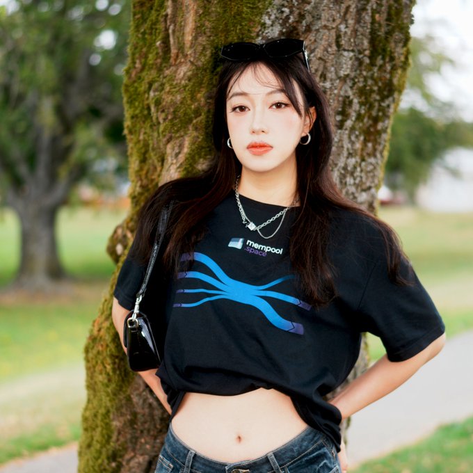 A young woman with long dark hair, sunglasses on her head, and silver jewelry, wearing a black crop top with a blue tree-like graphic and 'mempool space' logo, posing against a moss-covered tree in a park.