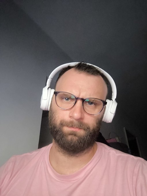 A selfie of a bearded man wearing glasses, white over-ear headphones, and a pink t-shirt against a dark background.