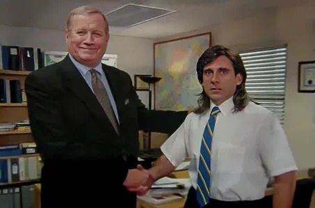 Two men shaking hands in an office, one older in a dark suit smiling and one younger with long hair in a white shirt and blue striped tie looking awkward.