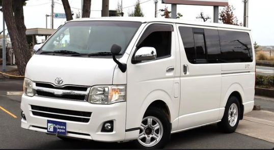 A white Toyota HiAce passenger van is parked outdoors, featuring the "Hokkaido Japan" logo on its front license plate area.