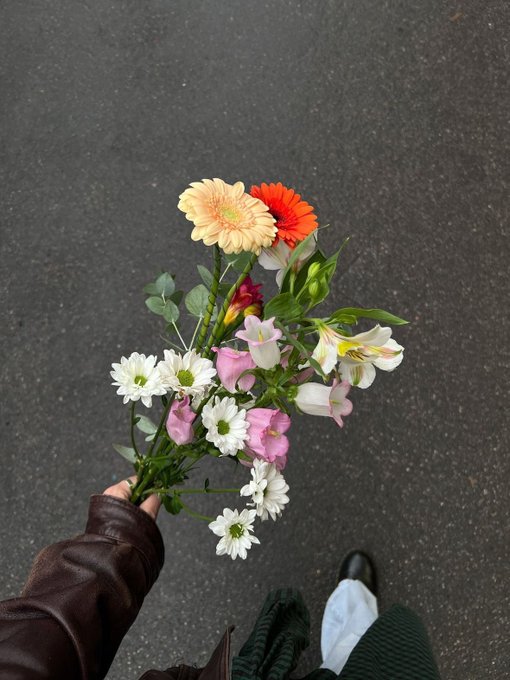A first-person view of a hand holding a colorful bouquet of flowers including white daisies, pink blossoms, orange gerbera, and alstroemeria over asphalt pavement.