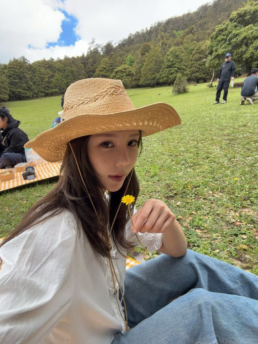 A young woman wearing a straw sun hat and white blouse sits on a grassy field holding a small yellow flower, with people relaxing on picnic blankets in the background. The caption reflects on Robinhood's $CASHCAT token's rapid rise to a $110M market cap, with the woman's peaceful outdoor scene contrasting the fast-paced crypto launch.