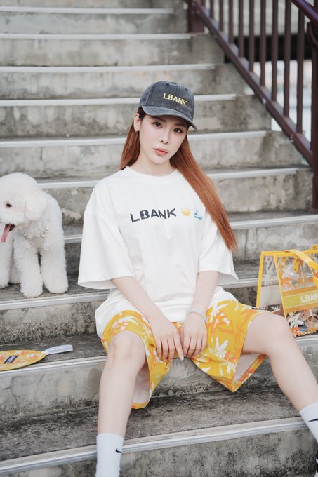 A young woman wearing an "LBANK" cap and matching white t-shirt sits on concrete steps beside a fluffy white dog and a yellow LBANK-themed bag, perfectly capturing the casual summer street style of the LBank Summer Mode campaign.