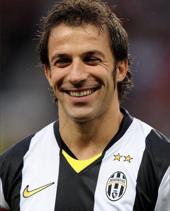 A smiling man with dark hair wearing a black and white striped Juventus football jersey with yellow accents and the team crest.
