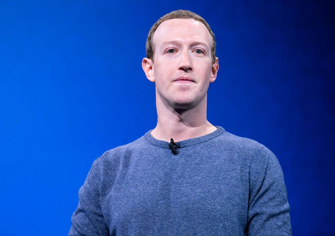 Mark Zuckerberg stands on stage wearing a gray sweater with a microphone attached, photographed against a blue background.