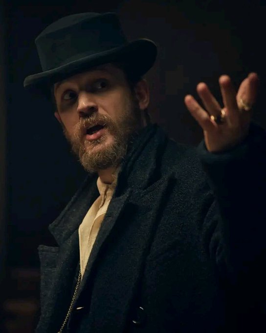 A man with a beard, wearing a black top hat and coat, looking intensely with his hand raised, revealing multiple rings.