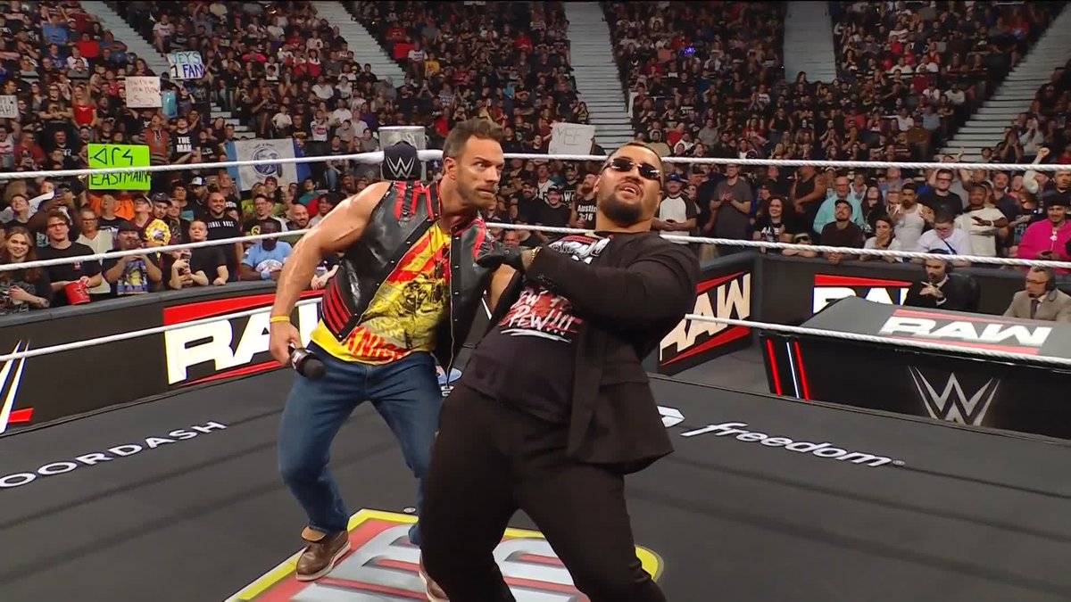 Two men are in a WWE wrestling ring, with one wearing a black vest over a yellow shirt and the other in a black shirt with sunglasses; the crowd and 'RAW' ring apron are visible in the background.