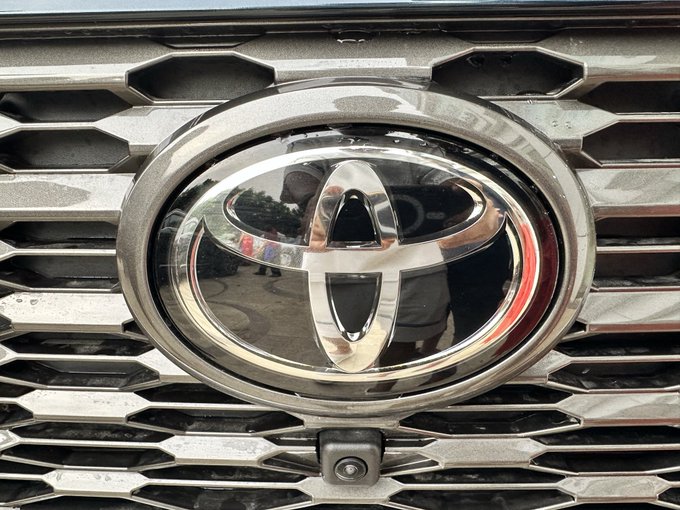 A close-up of a chrome Toyota emblem on a vehicle's front grille with a honeycomb pattern, with the photographer's reflection visible in the shiny surface.