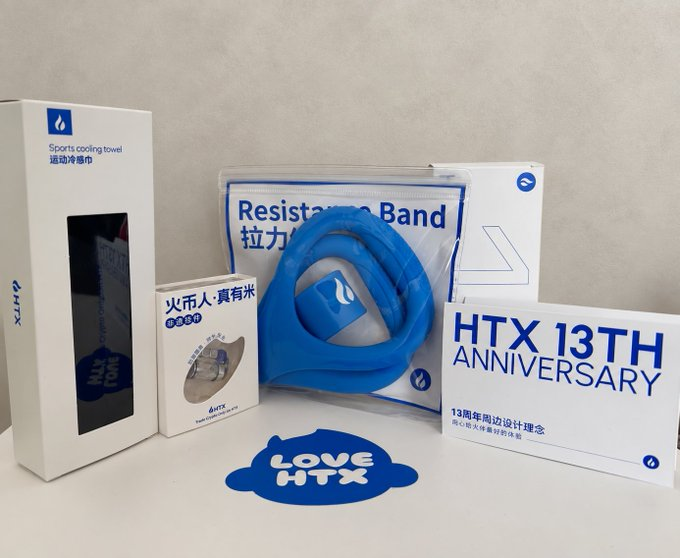 A promotional product display featuring a sports cooling towel box, resistance band, rice packaging, and 'HTX 13TH ANNIVERSARY' branded items with a 'LOVE HTX' logo.