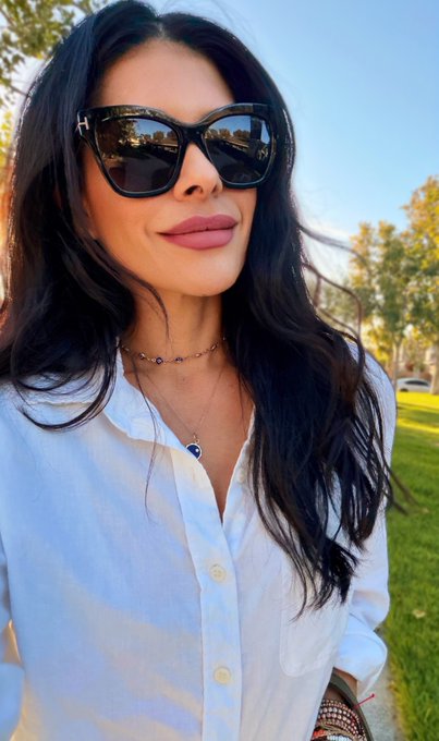 A woman with long dark hair wears oversized tortoiseshell sunglasses and a crisp white button-up shirt, accessorized with delicate necklaces, standing outdoors on a sunny day. The warm, confident pose pairs with the message of gratitude and quiet strength, reflecting a serene start to a Thursday morning.