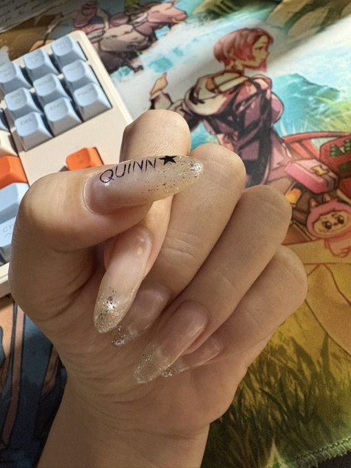 A hand displays long, glittery coffin-shaped nails with the name "QUINN" and a star painted on the middle finger, resting above a white keyboard and an anime-themed desk mat. The image aligns with the post's caption, which discusses how the long nails make typing painful, referencing @quipnetwork.