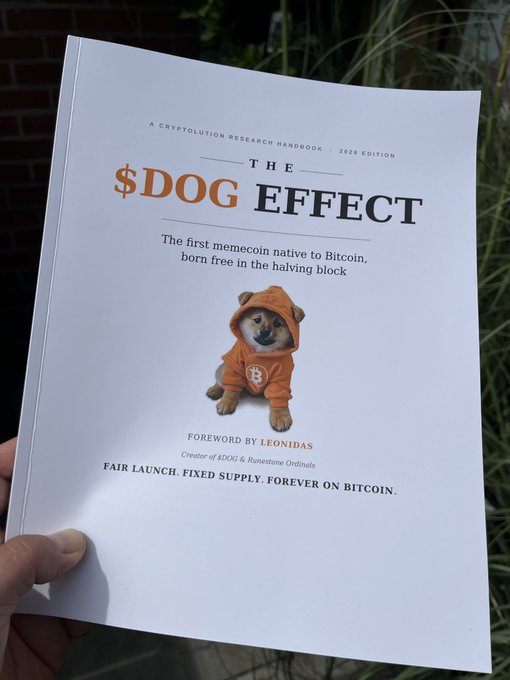 A hand holds a printed book titled '$DOG EFFECT' described as 'A Cryptolution Research Handbook · 2026 Edition' about a memecoin native to Bitcoin, featuring a Shiba Inu dog in an orange hoodie with a Bitcoin logo.
