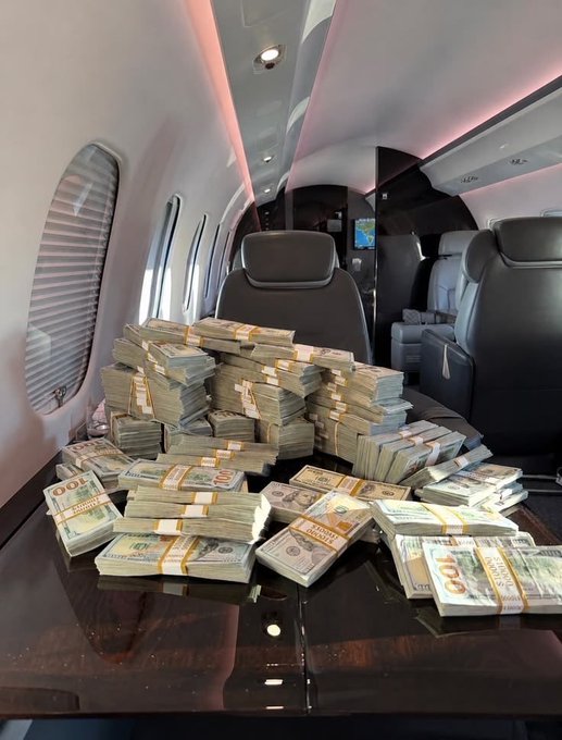 Stacks of bundled US hundred-dollar bills piled on a wooden table inside the cabin of a private jet.