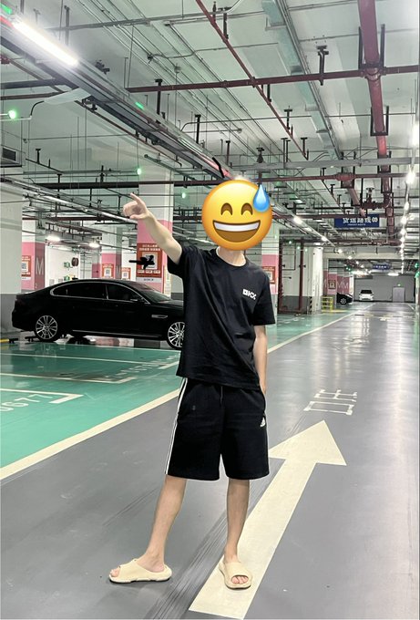 A young person wearing a black t-shirt, black shorts, and yellow slide sandals stands in a parking garage pointing upward, with a smiley face emoji covering their face, and a black car visible in the background.