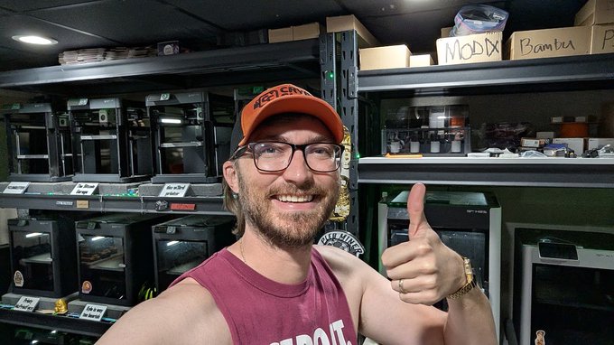 A bearded man wearing glasses and an orange 'ARIZONA' cap, giving a thumbs-up in a room filled with 3D printers on metal shelving, with boxes labeled 'MODX' and 'Bambu' visible on top.