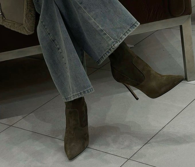 A close-up of a person sitting down, wearing light blue jeans and olive green suede pointed-toe Chelsea boots with stiletto heels, on a tiled floor.