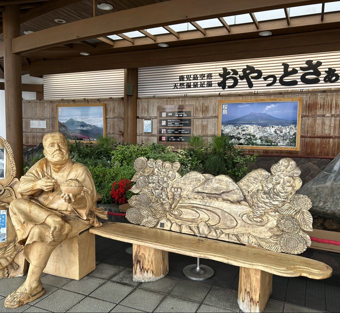 A wooden carved statue of a seated figure sits beside a long wooden bench with intricate floral carvings, displayed in what appears to be a Japanese rest area with Japanese signage including 'おやっとさぁ'.