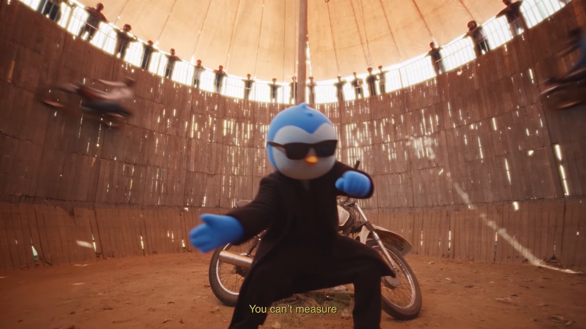 The same blue penguin mascot character wearing sunglasses and a black suit performs in front of a motorcycle inside a wooden 'Wall of Death' carnival ride, with the subtitle 'You can't measure' displayed.