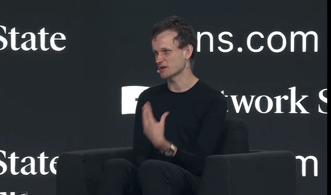 A man in a black long-sleeve shirt is speaking at a 'Network State' conference, seated on a dark couch in front of a backdrop displaying 'networkstate.com'.