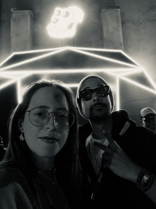 A black-and-white selfie of a woman with glasses and a man wearing sunglasses and a beanie making a peace sign, taken at night under a glowing neon skull sign.