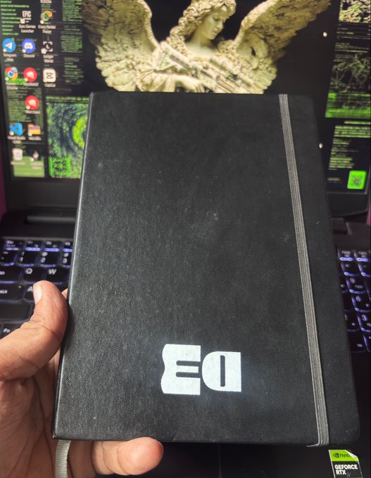 A hand holds a black notebook with a stylized "EO" logo in front of a laptop displaying a green angel wallpaper and code.