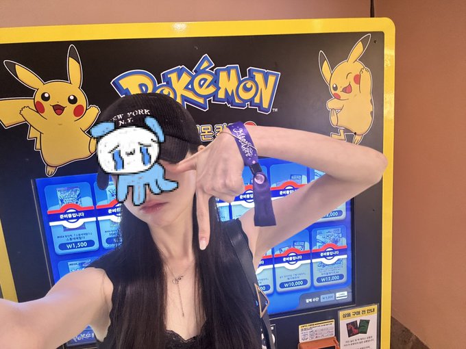 A young woman wearing a black 'NEW YORK N.Y.' cap and a purple lanyard poses in front of a Pokémon vending machine, making a peace sign; her face is obscured by a pixelated squid sticker.