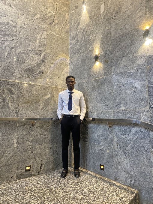 A young man in a white dress shirt, blue tie, and dark trousers stands against a gray marble wall in a corner with wall sconces.