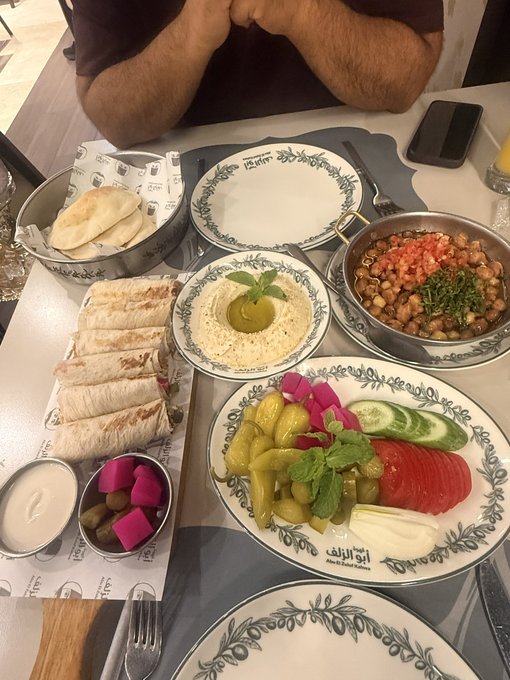 A spread of Middle Eastern food is laid out on a table including hummus with mint, fattoush salad, pickled vegetables, pita bread, and a chickpea dish, with Arabic text visible on one plate.