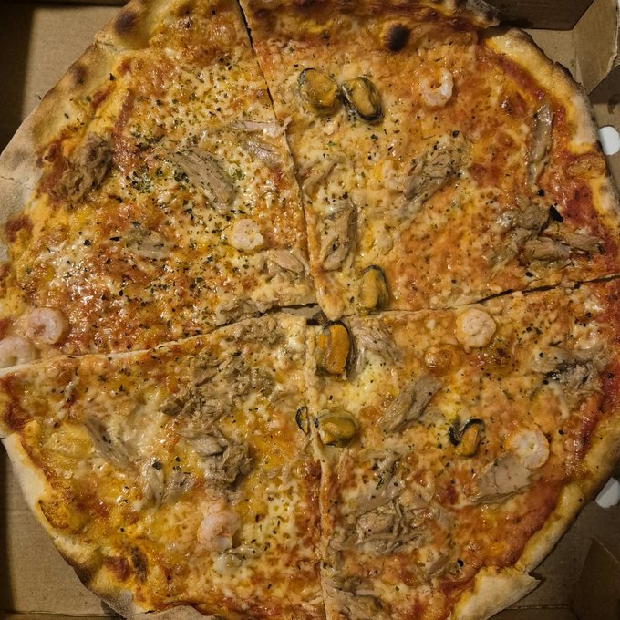 A close-up overhead view of a seafood pizza topped with cheese, shrimp, mussels, and herbs, sliced into eight pieces in a cardboard box.