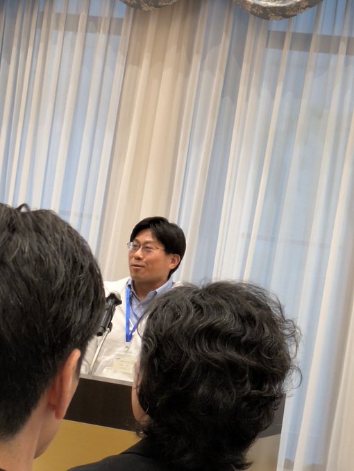 A man in a white shirt with a lanyard speaks at a microphone during what appears to be a conference or panel discussion, viewed from behind audience members. The setting features light-colored curtains in the background, consistent with the accompanying post introducing the speaker as the Stablecoin Section Manager of Adomi Cryptocurrency Assets.