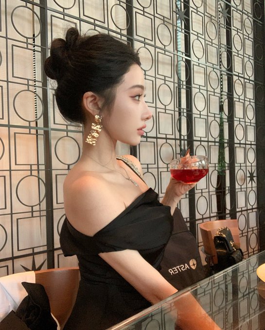 A woman in an elegant black off-shoulder dress poses in profile against a geometric-patterned wall, holding a glass of red cocktail, with a handbag displaying the "ASTER" brand visible on her lap.

The Aster-branded bag in the image directly ties to the post's theme about being a devoted holder of the Aster cryptocurrency, symbolizing loyalty and a stylish, sophisticated approach to long-term holding without checking or selling.