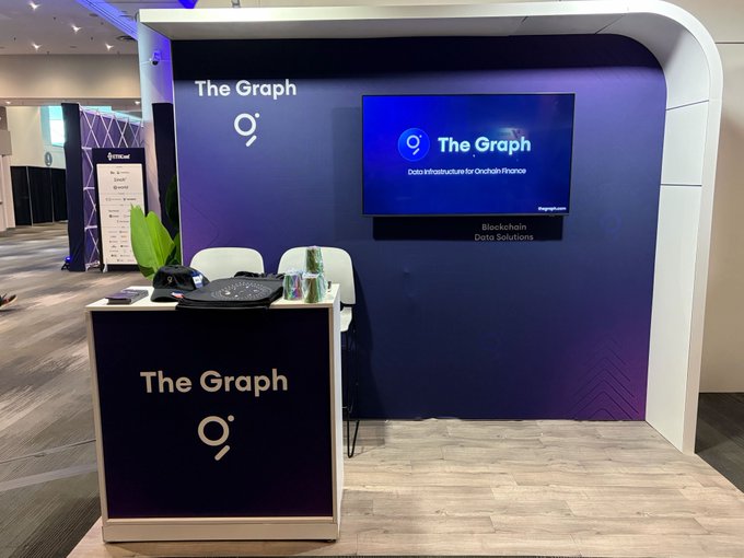 The image shows a booth display with a white countertop set up at an expo or trade show. On the counter, there are two chairs placed facing each other, next to a television screen displaying The Graph logo and information about the event. A person is also present in the scene near the TV. In addition to these elements, there's another smaller TV visible on the left side of the image. There is a phone positioned towards the center-left corner of the booth display.