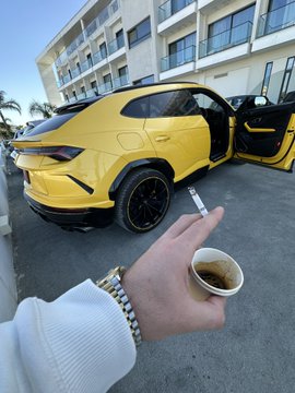 A vibrant yellow Lamborghini Urus with its door open is parked outside, as a person's hand holding a small coffee cup extends into the frame, perfectly capturing the laid-back luxury "vibe check" moment.
