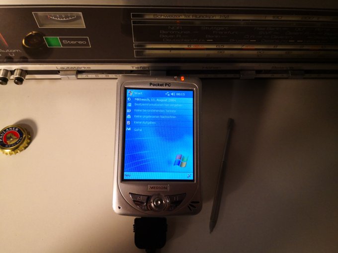 A Medion Pocket PC device displaying a Windows-style Start menu sits on a desk beneath a Schneider IV Receiver radio, with a bottle cap and stylus nearby.