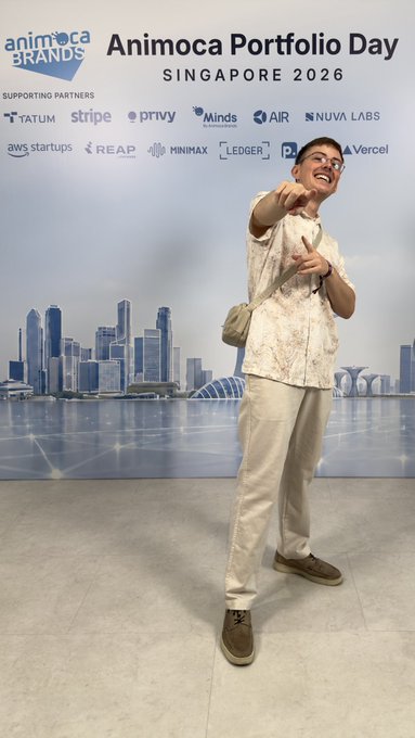 A man in a patterned shirt and light pants poses and points at the camera at the 'Animoca Portfolio Day Singapore 2026' event, with logos for supporting partners like TATUM, stripe, privy, AIR, NUVA LABS, aws startup, MINIMAX, LEDGER, and Vercel visible on the backdrop.