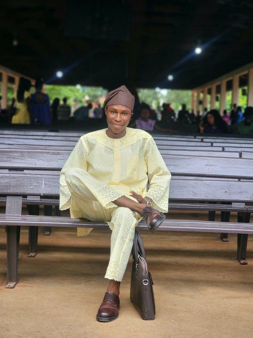 A smiling young man in a pale yellow traditional embroidered outfit and brown cap sits on a wooden bench inside a covered hall, with a dark leather bag placed on the floor beside him.
