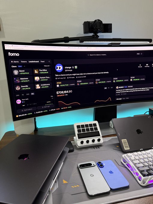 A workspace featuring a curved monitor displaying the Fomo trading platform with a portfolio showing $708,164.90, alongside two MacBook Pros, a Pixel and iPhone, and a mechanical keyboard. The image reflects the post's cautionary message about meme coin trading, contrasting an impressive-looking portfolio balance with the risky, community-driven nature of such investments that the post warns against.