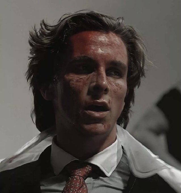 A cinematic portrait of a young man with messy dark hair, wearing a white shirt and patterned tie, his face marked with blood and bruises.