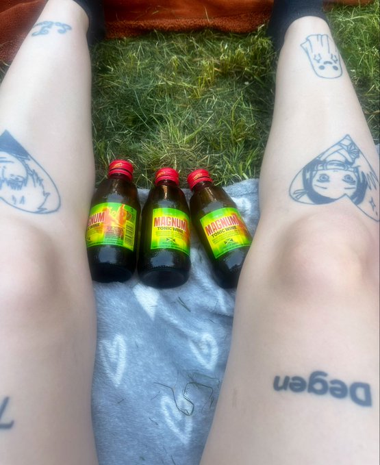 Three small bottles of Magnum tonic wine sit on a blanket between a person's tattooed legs, which feature anime-style designs and the word "vegan."